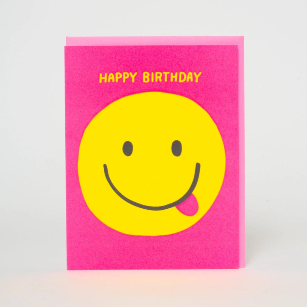 Birthday card with a happy face with their tongue sticking out on a bright pink background. Yellow Happy Birthday printed above. A pink envelope is partially visible behind the card.  Blank inside