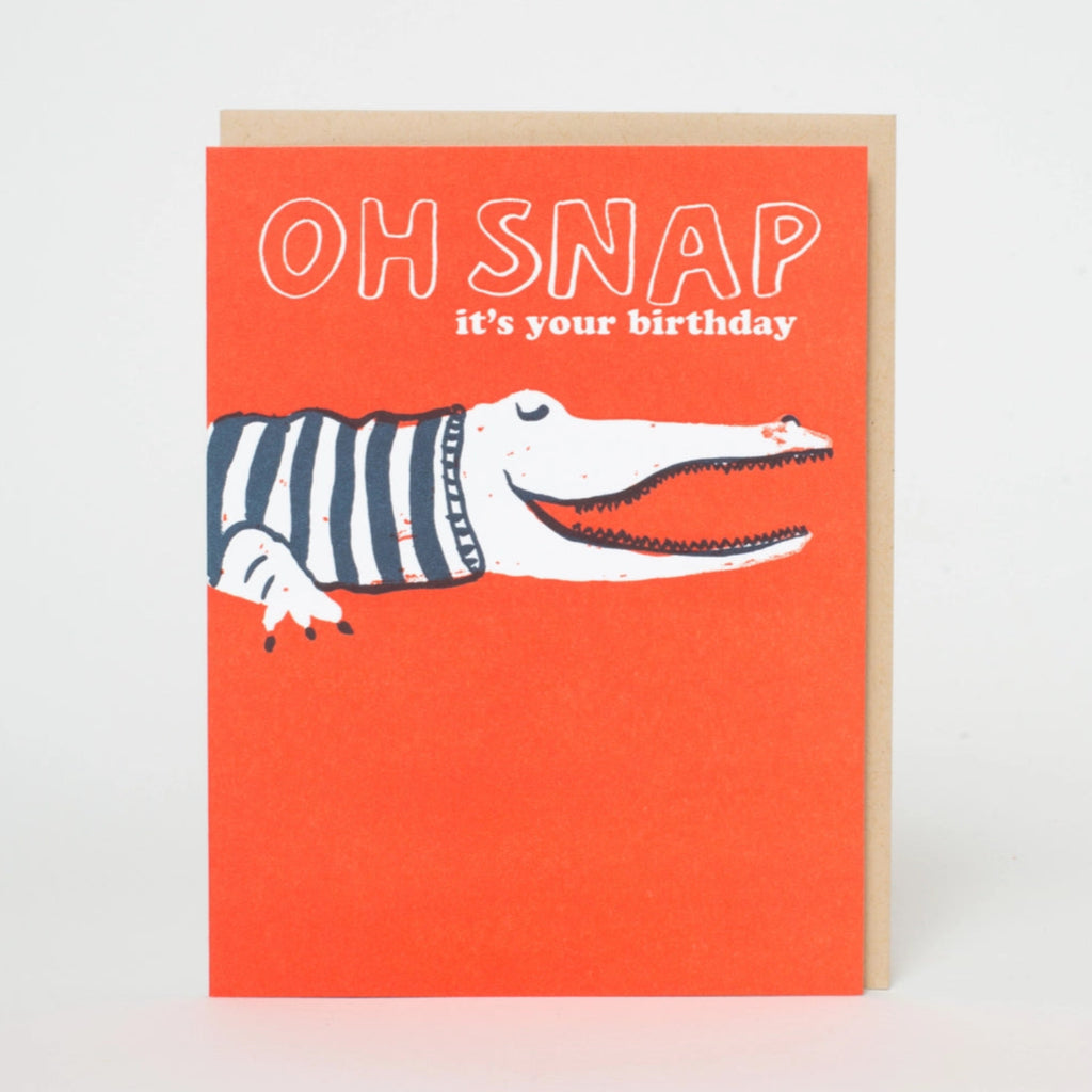A birthday card with an illustration of an alligator wearing a striped shirt, with the text 'OH SNAP it's your Birthday' on an orange background. A envelope is partially visible behind the card.  Blank inside