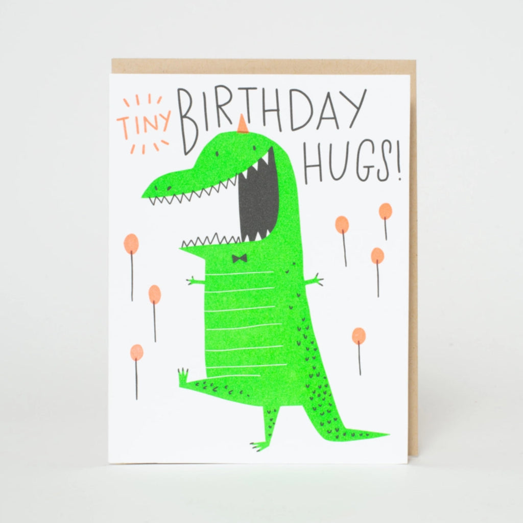 A birthday card with an illustration of a happy green dinosaur with the text 'Tiny Birthday Hugs' on a white background. A envelope is partially visible behind the card.  Blank inside